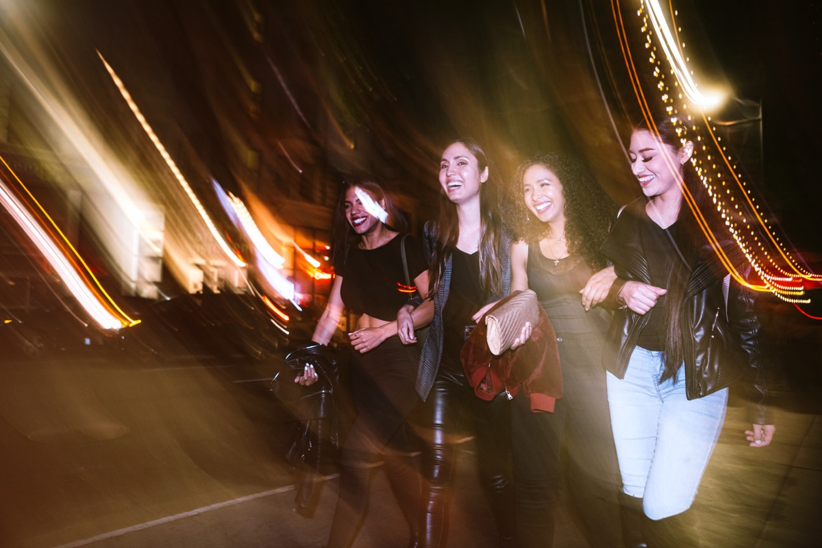 Four friends walking arm in arm through city lights at night after the crawl